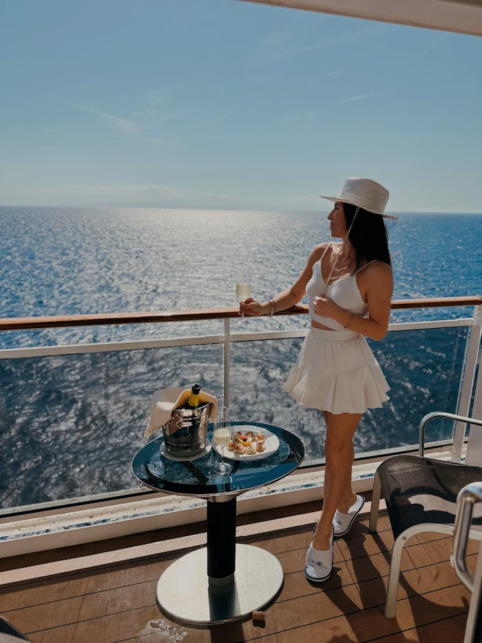 Woman in white attire holding a drink on a cruise ship deck, gazing at the ocean.